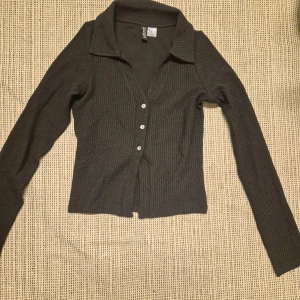 Svart ribbad kofta från H&M Divided - Snygg svart ribbad kofta från H&M Divided med knappar framtill. Perfekt för lager-på-lager och att hålla sig varm under kyliga dagar. Den har en klassisk krage och långa ärmar.