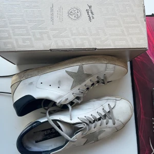 Golden Goose vita sneakers - Säljer ett par stilrena vita sneakers från Golden Goose med den ikoniska stjärndetaljen i silver på sidan. Skorna har en mörk blå häl och snörning framtill. Perfekta för en avslappnad men trendig look. Köpta nya från Golden goose hemsida för 5400 endast använda i 3 månader mitt pris 3500