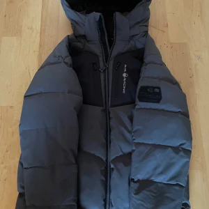 Grå dunjacka från Sail Racing - Snygg och varm grå dunjacka från Sail Racing med huva och dragkedja. Perfekt för kalla dagar med sin pufferdesign och stilrena look. Jackan har flera praktiska fickor och en logotyp på bröstet.