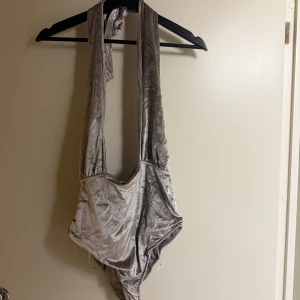 Grå sammet bodysuit från Bik Bok - Snygg grå bodysuit i mjukt sammetstyg från Bik Bok. Den har en djup urringning och breda axelband som knyts i nacken. Perfekt för en stilren look vid poolen eller stranden.