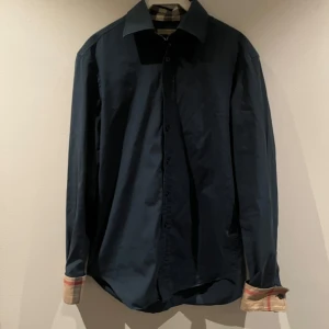 Mörkblå skjorta från Burberry - Snygg mörkblå skjorta från Burberry med klassiskt rutigt mönster på insidan av kragen och ärmsluten. Skjortan är i bra skick och sparsamt använd. Vid fler frågor hör gärna av dig. Vid snabb affär kan priset diskuteras.
