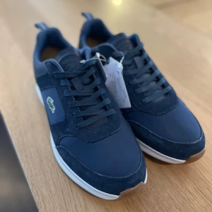 Mörkblå sneakers från Lacoste - Snygga mörkblå sneakers från Lacoste med vit sula och klassisk krokodillogga på sidan. Skorna är slip-on alltså du behöver inte slösa tid på att knyta skorna, perfekt för vardagsbruk. Yttersulan är i gummi för bra grepp. De är i storlek 42 men är väldigt små för den storleken!