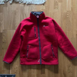 Röd fleecejacka från Geographical Norway - Säljer en röd fleecejacka från Geographical Norway med dragkedja och flera praktiska fickor. Jackan har en hög krage och broderad logga på ärmen. Perfekt för kyliga dagar!