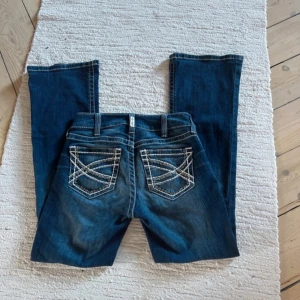 Lågmidjade bootcut jeans  - Snygga blå bootcut jeans med vita broderade detaljer på bakfickorna. De har en klassisk femficksmodell och en bekväm passform. Midjemåttet rakt över: 37 cm      Innerbenslängden: 78cm                                      Jag är runt 164 cm                                         Grenhöjd: 19cm 