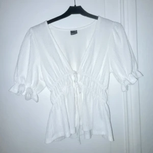 Vit blus med knytning - Säljer en söt vit blus med v-ringning och knytning i midjan. Blusen har korta puffärmar med volangdetaljer och en luftig passform. Perfekt för vår och sommar!
