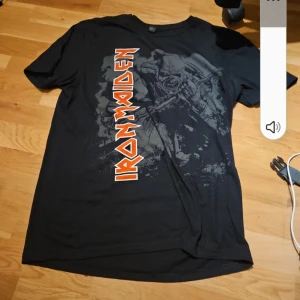 Svart Iron Maiden t-shirt - Cool svart t-shirt med Iron Maiden-tryck i rött och vitt. Perfekt för fans av bandet. T-shirten har korta ärmar och ett stort tryck på framsidan.