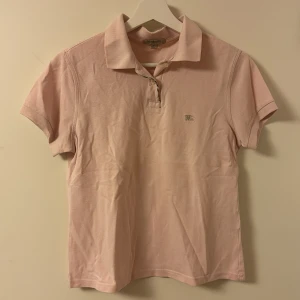 Burrberry Pikée Babyrosa - Säljer denna fina burrberry skjortan i en riktigt stilren färg, en lite ljusare ton av rosa. Pikén är i storlek M men är liten i storleken så passar ungefär S/XS. Skicket är 8/10 då det finns ett pyttelitet hål vid en av knapparna som it syns.