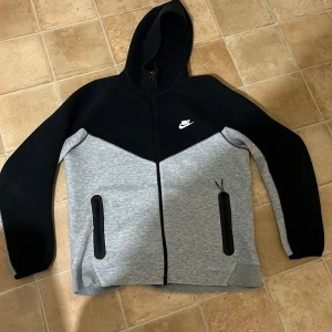 Grå och svart hoodie från Nike - Säljer en snygg och bekväm grå och svart hoodie från Nike. Den har en dragkedja framtill och två praktiska fickor med dragkedjor. Perfekt för både träning och vardag. Hoodien är i nyskick och har en klassisk Nike-logga på bröstet.