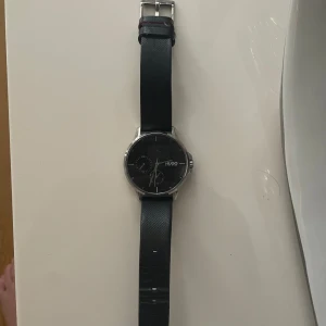 Svart klocka från Hugo - Säljer en stilren svart klocka från Hugo Boss med en elegant design. Klockan har en svart urtavla och ett matchande svart armband i skinn. Perfekt för både vardag och fest! Original pris 1996kr