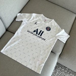 Vit PSG fotbollströja - Säljer en vit PSG fotbollströja med korta ärmar. Tröjan har 'Accor Live Limitless' tryckt på framsidan och 'Visit Rwanda' på baksidan. Den har ett diskret mönster med små texttryck över hela tyget. Perfekt för alla PSG-fans!