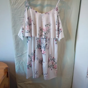 Blommig sommarklänning från Shein - Säljer en superfin blommig klänning från Sheilay, perfekt för sommardagar! Den är vit med ett vackert blommönster i rosa och grönt. Klänningen har en volangdetalj upptill och är ärmlös med smala axelband. Materialet känns lätt och luftigt, perfekt för varma dagar. Prislappen kvar 