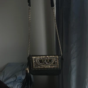 Svart axelväska med nitar - Snygg svart axelväska med kedjeaxelrem och coola nitar på framsidan. Väskan har en dekorativ fransdetalj på sidan som ger den extra edge. Perfekt för att ge din outfit en rockig touch.