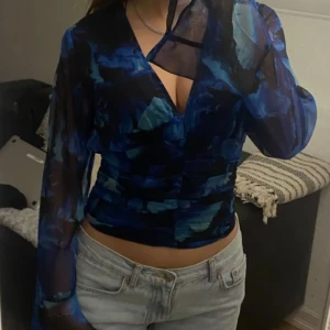 Blå blus med blommigt mönster - Snygg blå blus med blommigt mönster och transparenta långa ärmar. Blusen har en djup v-ringning och är perfekt för en stilfull look.