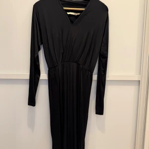 Svart långklänning från House of Lola - Elegant svart långklänning från House of Lola i mjukt och stretchigt material. För mig som är 1,63 cm så räcker den ca 10 cm nedanför knäna. Klänningen har en v-ringad hals, långa ärmar och en mjuk resår i midjan. perfekt för en stilren look. Den består av 91% viskos vilket ger den en glansig yta och fint fall. Klänningen är lika snygg med ett par sneakers som med klackskor. Endast använd 2 gånger så i jättefint skick!