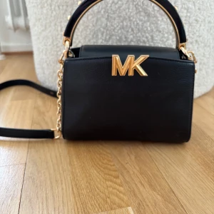Svart handväska med gulddetaljer - Snygg svart handväska från Michael Kors med guldfärgad logotyp och detaljer. Väskan har en elegant kedja och ett praktiskt handtag. Perfekt för att ge din outfit en lyxig touch.