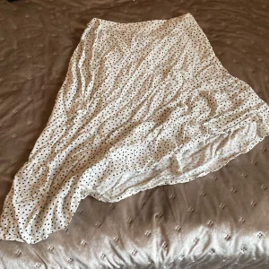 Vit prickig klänning med knytband - Charmig vit klänning med svarta prickar och knytband i midjan. Perfekt för en somrig look. Klänningen har en luftig passform och är tillverkad i ett lätt material. Den är längre på ena sidan och kortare på andra. Det är resår band på midjan så kan passa oavsett storlek. Den är väldigt luftig och sitter inte så tajt. 