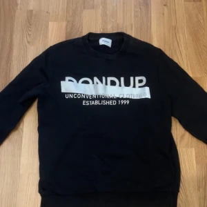 Svart sweatshirt från Dondup - Säljer en svart sweatshirt från Dondup med texten 'Unconventional Clothing Established 1999' i silver på framsidan. Tröjan har en rund halsringning och långa ärmar. Perfekt för en avslappnad stil. 