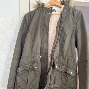 Grön jacka  - Säljer en grön jacka med fuskpäls på huvan. Jackan har dragkedja och knappar framtill samt stora fickor med knäppning. Insidan är fodrad med mjukt, beige foder för extra värme. Jackan är sparsamt använd och är i mycket bra  skick.