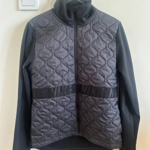 Svart quiltad jacka från Nike - Snygg svart quiltad jacka från Nike med dragkedja och hög krage. Jackan har ett stilrent mönster och är perfekt för löpning eller vardagsbruk. Den har långa ärmar och en diskret Nike-logga på bröstet.