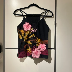 Svart linne med blommigt tryck - Snyggt svart linne med tunna axelband och färgglatt blommigt tryck i rosa och gult. Linnet har en avslappnad passform och är tillverkat i ett mjukt material.