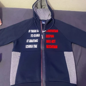Mörkblå hoodie från Arma - Säljer en mörkblå hoodie från Arma med gråa detaljer och texttryck i vitt och rött. Hoodien har en dragkedja framtill och en justerbar huva. Perfekt för en avslappnad stil. Helt ny utan prislapp och helt oanvänd. 