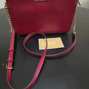 Vinröd Jet Set från Michael Kors oanvänd - Snygg vinröd axelväska från Michael Kors i skinn med guldfärgade detaljer och justerbar axelrem. Väskan har en dragkedja upptill och ett rymligt foder med plats för dina essentials. Perfekt för att ge din outfit en lyxig touch.