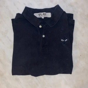 Svart pikétröja från Comme des Garçons - Säljer en L svart pikétröja från Comme des Garçons med det ikoniska hjärtemärket på bröstet. Tröjan har en klassisk krage med knappar. Perfekt för en minimalistisk look. Den passar både L och M. nypris runt 900kr