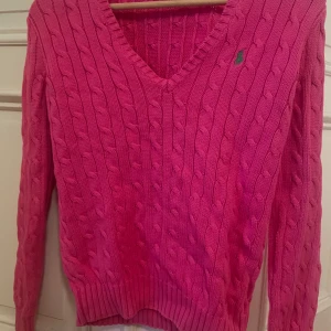 Rosa stickad tröja från Ralph Lauren - Snygg rosa stickad tröja från Ralph Lauren med kabelstickat mönster och v-ringning. Tröjan har långa ärmar och en liten logga på bröstet. Perfekt för en stilren och bekväm look.