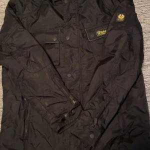 Svart jacka från Belstaff - Säljer en svart jacka från Belstaff med dragkedja och knappar. Jackan har en klassisk design med flera fickor och ett rutigt foder. Perfekt för höst och vår. nypris ligger på runt 6000kr. pris kan diskuteras