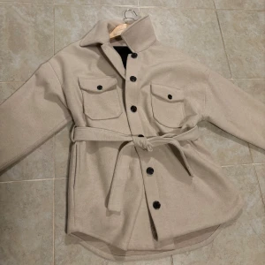 Beige jacka från NLY Trend - Snygg beige jacka från NLY Trend med knappar och bälte i midjan. Jackan har två bröstfickor med knappar och en klassisk krage. Fint skick och säljer pga att ja ej använder.