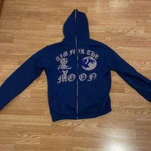 Blå hoodie med dragkedja och text - Säljer en blå hoodie med dragkedja från Aim for the Moon. Den har ett unikt tryck med texten 'Aim for the Moon' och en cool design på framsidan. Perfekt för en avslappnad stil.