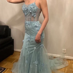 Ljusblå långklänning med blommor - Elegant ljusblå långklänning med blommiga detaljer och snörning i ryggen. Klänningen har en vacker transparent överdel med blommor och pärlor, samt en flytande kjol i tyll. Perfekt för en speciell kväll! Bal,bröllop,dop osånt. Storlek 4.  Xs blir de.  Passar även s    Aldrig använd 