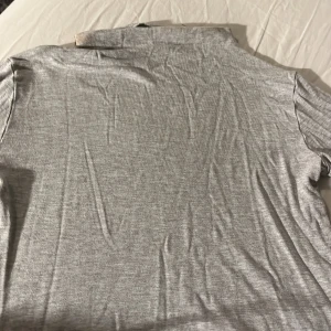 Grå pikétröja - Säljer en stilren grå pikétröja i mjukt material. Perfekt för en avslappnad men ändå snygg look. Tröjan har en klassisk krage och korta ärmar.