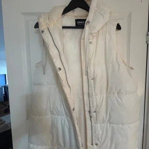 Vit dunväst från ONLY - Säljer en snygg vit dunväst från ONLY. Västen har en dragkedja framtill och är perfekt för kyligare dagar. Den är stilren och lätt att matcha med olika outfits. Har huva, är medel lång. Köpt på Vera Moda för 400kr, säljer 200kr.