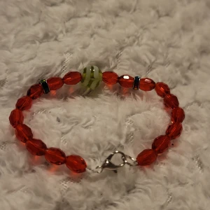 Rött armband med pärlor - Charmigt armband med röda facetterade pärlor och en unik grön pärla med mönster. Armbandet har en praktisk metallspänne för enkel användning. Perfekt för att lägga till en färgklick till din outfit.