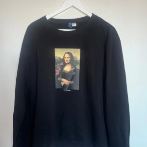 Svart sweatshirt med Mona Lisa-tryck - Snygg svart sweatshirt med ett stort tryck av Mona Lisa på framsidan. Tröjan har långa ärmar och en rund halsringning. Perfekt för dig som vill ha en konstnärlig touch i garderoben. Från H&M i stl XL