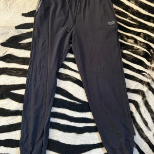 Svarta mjukisbyxor från Hugo Boss - Snygga svarta mjukisbyxor från Hugo Boss med dragsko i midjan för justerbar passform. Perfekta för en avslappnad stil. Byxorna har en diskret logga på sidan och är tillverkade i ett mjukt material för extra komfort. Obs. lite slitet skick vid fickorna som går att fixa väldigt lätt! 