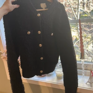 Svart kofta med knappar - Snygg svart kofta med glittriga knappar och en elegant design. Tröjan har långa ärmar och en klassisk passform. Perfekt för att ge en stilren look till din outfit.