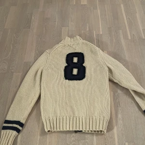 Beige stickad tröja med nummerdetalj - Snygg beige stickad tröja med dragkedja och mörkblå detaljer. Tröjan har ett stort nummer 8 på ryggen och ribbade muddar med ränder på ärmarna. Perfekt för en avslappnad stil.