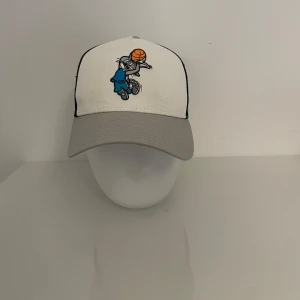 Keps  - Snygg keps i vitt och grått med ett coolt broderat basketmotiv på framsidan. Baksidan är i svart meshmaterial för bra ventilation. Perfekt för sportiga tillfällen eller som en stilren accessoar.