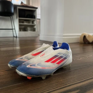 Adidas F50 fotbollsskor - Snygga Adidas F50 fotbollsskor i vitt med röda och blå detaljer. Skorna har en strömlinjeformad design med en låg profil och är perfekta för snabba rörelser på planen.
