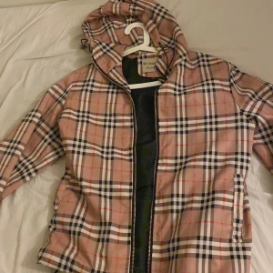 Rutig jacka från Burberry - Säljer en snygg jacka från Burberry i klassiskt mönster. Jackan har en dragkedja framtill och en huva. Perfekt för att ge din outfit en stilren touch.
