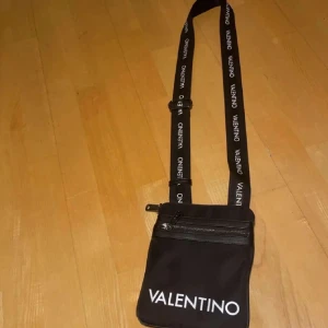 Svart axelväska från Valentino - Snygg svart axelväska från Valentino med justerbar axelrem och dragkedja. Väskan har en minimalistisk design med logotypen tydligt synlig på framsidan.