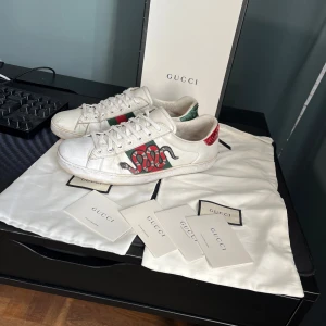 Gucci ace skor - Snygga vita sneakers från Gucci med broderade detaljer i rött och grönt på sidan. Skorna har klassisk snörning och den ikoniska Gucci-randen i grönt och rött. Perfekta för att ge din outfit en lyxig touch. Skicket får man själv bedöma efter bilderna! Skriv vid minsta fundering, själv klart äkta och allt og till kommer