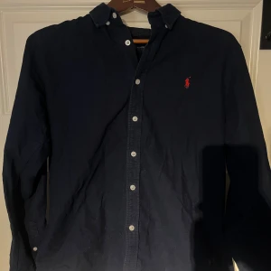 Linné skjorta från Ralph Lauren - Säljer en stilren och tidlös Linné-skjorta från Ralph Lauren i storlek L (Slim Fit). Passformen gör att den även sitter bra på dig som vanligtvis bär M. Den är självklart äkta och nypris ligger på 1 995kr  ✅ Mörkblå med klassiska detaljer ✅ Ikoniska broderade loggan i rött ✅ Knappar med Ralph Lauren-gravyr ✅ Mycket bra skick – sparsamt använd  Perfekt för både vardag och uppklädda tillfällen! Skicka ett meddelande vid intresse.