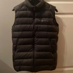 Svart dunväst från Sail Racing - Säljer en svart dunväst från Sail Racing. Västen har en dragkedja framtill och är quiltad för extra värme. Perfekt för kyligare dagar och har en stilren design med logotyp på bröstet och ryggen. Den har två fickor där fram och en lite mindre inne ficka.