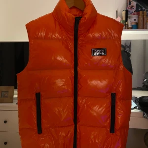 Orange dunväst från DSQ2 - Säljer en cool orange dunväst från DSQ2 med hög krage och dragkedja framtill. Västen har två svarta dragkedjefickor och en stor logga på bröstet samt en patch på ryggen. Perfekt för att sticka ut i mängden!
