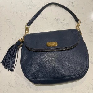 Mörkblå axelväska från Michael Kors - Snygg mörkblå axelväska i skinn från Michael Kors med gulddetaljer. Väskan har en avtagbar axelrem och en dekorativ tofs. Insidan är fodrad med logotypmönster och har flera fack, inklusive ett med dragkedja. Perfekt för att hålla ordning på dina saker med stil.