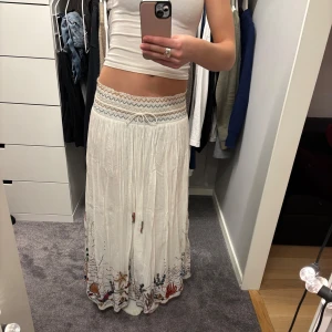 Vit långkjol med broderier - Säljer en vacker vit långkjol från zara med färgglada broderier längst ner. Kjolen har en elastisk midja med ett mönstrat band och snörning framtill. Så snygg till sommaren!!!🩷💛