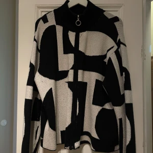 Svartvit mönstrad cardigan från NA-KD - Säljer en cool svartvit cardigan från NA-KD med ett unikt grafiskt mönster. Den har en dragkedja framtill och en hög krage med en ringdetalj. Perfekt för att ge din outfit en edgy touch. Något nopprig därav det billiga priset, men går att använda noppmaskin för att få bort